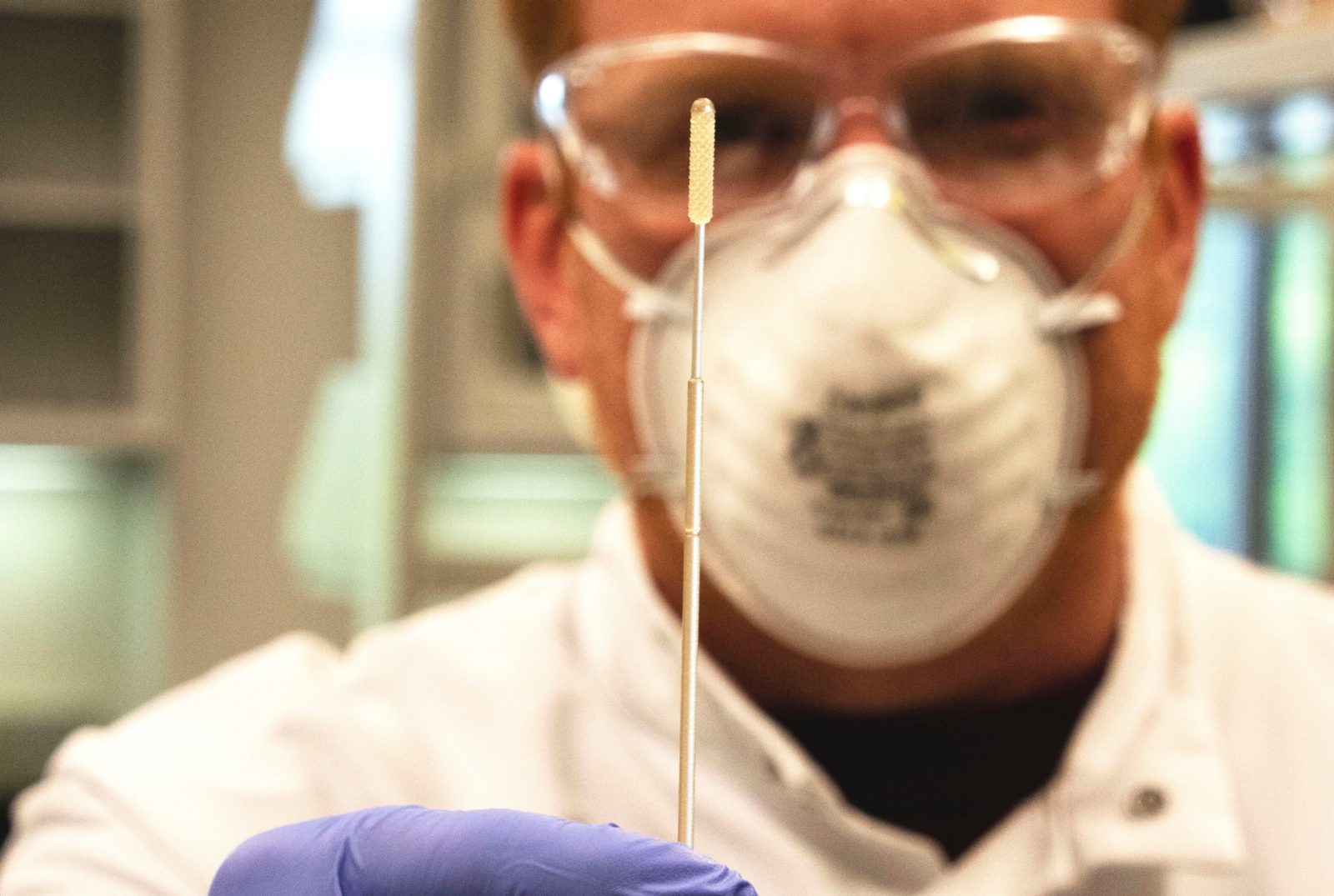 Marc Michel, an associate professor in the Department of Geosciences, holds up a nasopharyngeal swab that he 3D printed in a lab at Steger Hall. Michel, out of focus, is wearing gloves, a PPE mask, and googles in accordance with safety standards.