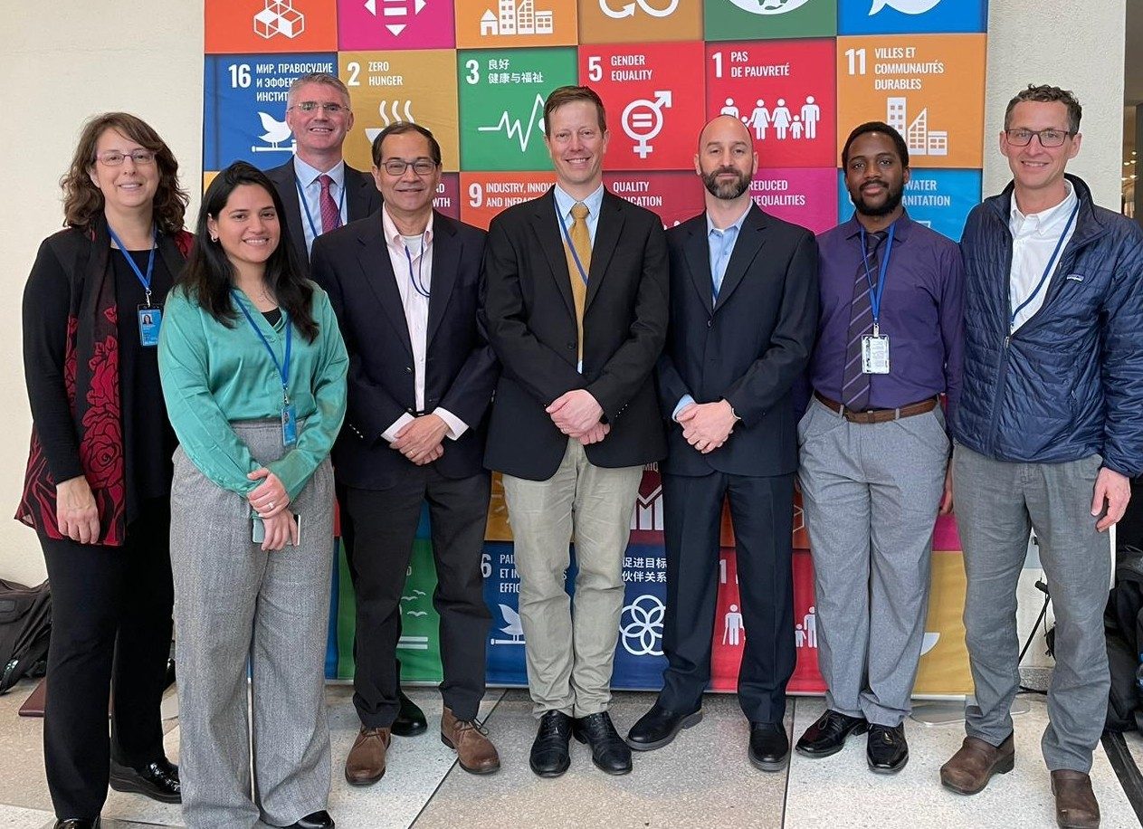 Eight scholars stand in front of a display board at the UN Water Conference